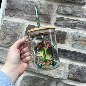 May include: A glass mug with a bamboo lid and a green straw. The mug has a graphic of a round door with a green door and a brick frame. The text on the mug reads "There and Back Again A Hobbit's Tale".