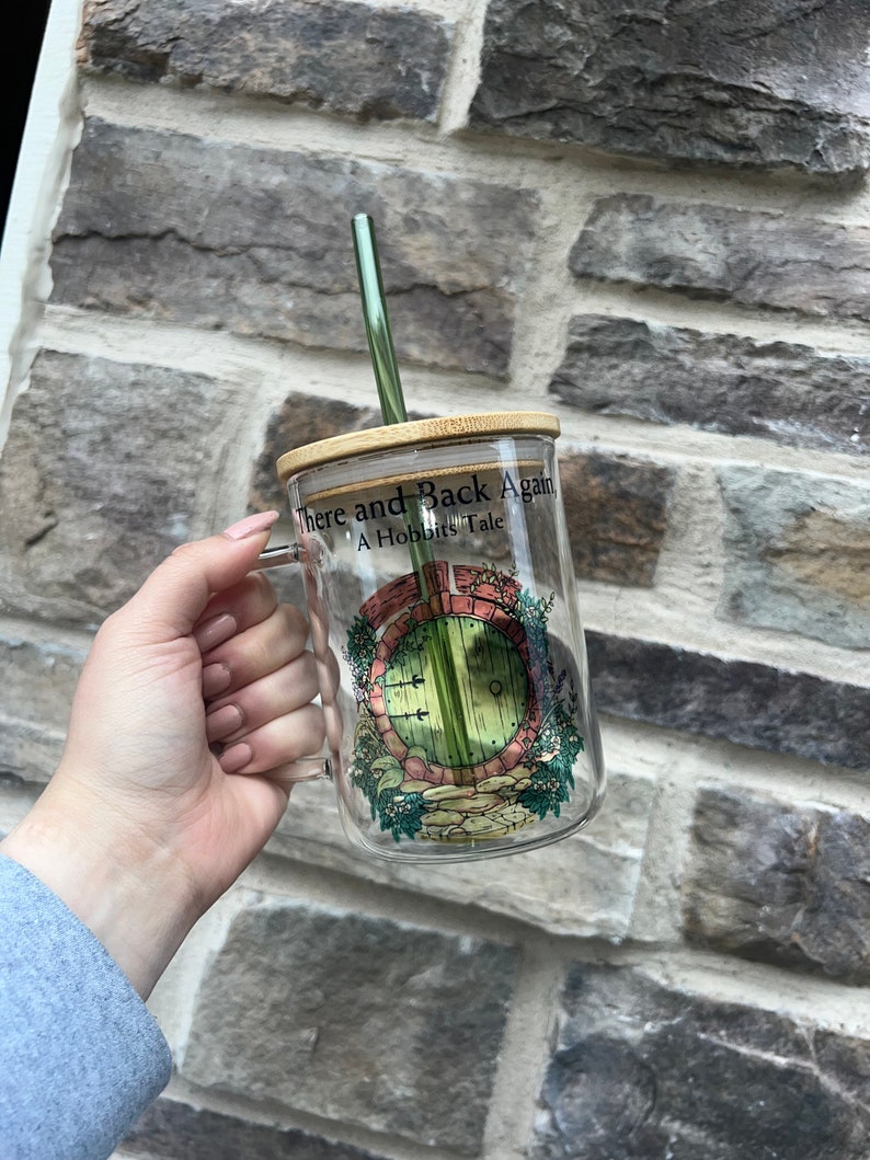 May include: A glass mug with a bamboo lid and a green straw. The mug has a graphic of a round door with a green door and a brick frame. The text on the mug reads "There and Back Again A Hobbit's Tale".