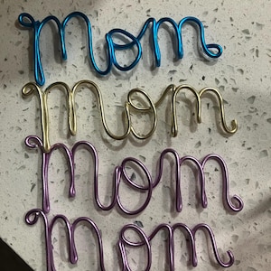 Puede incluir: Tres letreros de alambre con la palabra "Mamá" en azul, dorado y morado. La palabra "Mamá" está escrita en un estilo cursivo, con cada letra conectada. Los letreros están hechos de un material metálico brillante.