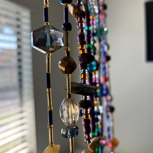 May include: Close-up of beaded strands with various shapes and colors. The strands feature faceted crystal, gold-colored beads, and a mix of other colorful beads. The background is blurred, with a window and blinds visible.