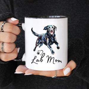 May include: White ceramic mug with a black rim and handle. The mug features a watercolour illustration of a black Labrador Retriever with a paw print and heart design. The text "Lab Mom" is written in black cursive below the dog.
