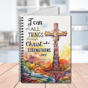May include: A spiral bound notebook with a colorful watercolor design featuring a cross and the quote "I can do all things through Christ who strengthens me" from Philippians 4:13.