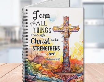 Op de afbeelding: Een spiraalgebonden notitieboek met een kleurrijk aquarelontwerp met een kruis en de quote "Ik kan alles door Christus, die mij kracht geeft" uit Filippenzen 4:13.