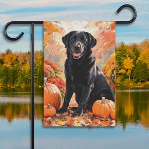 May include: A rectangular garden flag featuring a black Labrador retriever sitting among orange pumpkins and fallen autumn leaves. The background displays a watercolor-style forest with warm fall colors, all suspended from a black metal stand.