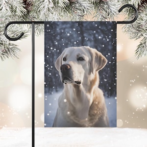May include: A white Labrador Retriever dog stands in a snowy winter scene. The dog is looking to the left of the image. The background is a blurry forest with falling snow.