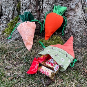May include: Three carrot-shaped fabric bags with green tops. Two bags are empty and one bag is filled with three Twix candy bars. The bags are sitting on a bed of green grass.