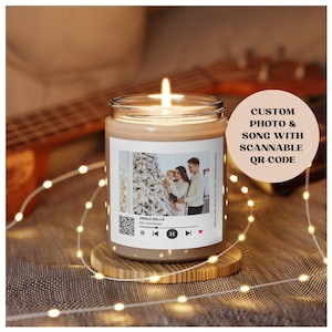 Puede incluir: Una vela marrón en un tarro de vidrio transparente con una etiqueta blanca. La etiqueta presenta una foto de una familia frente a un árbol de Navidad, un código QR y el texto "Jingle Bells - The Monkees".