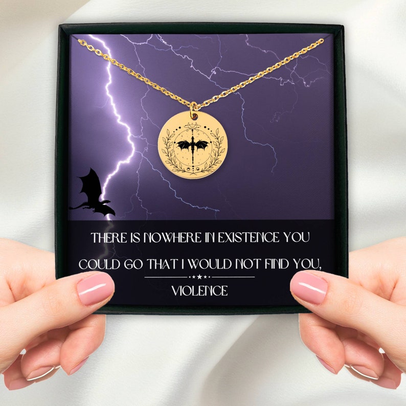 Peut inclure: Un collier en or avec un pendentif rond repr&eacute;sentant une silhouette de dragon noir &agrave; l'int&eacute;rieur d'une couronne. Le pendentif est dans une bo&icirc;te avec un fond violet et un motif d'&eacute;clair. Le texte sur la bo&icirc;te est : "THERE IS NOWHERE IN EXISTENCE YOU COULD GO THAT I WOULD NOT FIND YOU, VIOLENCE."