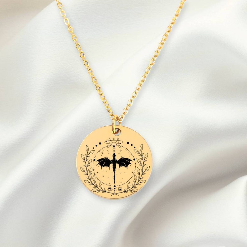 Peut inclure: Collier en cha&icirc;ne dor&eacute;e avec un pendentif rond repr&eacute;sentant une silhouette noire de dragon aux ailes d&eacute;ploy&eacute;es, entour&eacute;e d'un cercle de feuilles et de symboles c&eacute;lestes.