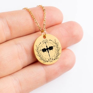 Peut inclure: Collier en cha&icirc;ne dor&eacute;e avec un pendentif rond repr&eacute;sentant un dragon noir aux ailes d&eacute;ploy&eacute;es, entour&eacute; d'une couronne de feuilles et de symboles c&eacute;lestes.