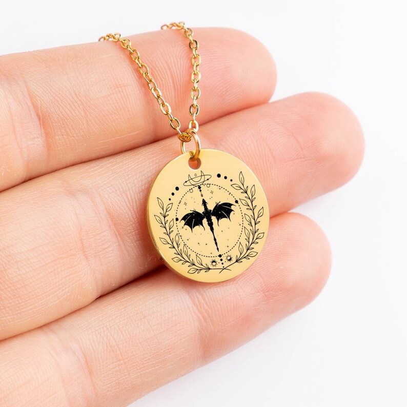 Peut inclure: Collier en cha&icirc;ne dor&eacute;e avec un pendentif rond repr&eacute;sentant un dragon noir aux ailes d&eacute;ploy&eacute;es, entour&eacute; d'une couronne de feuilles et de symboles c&eacute;lestes.