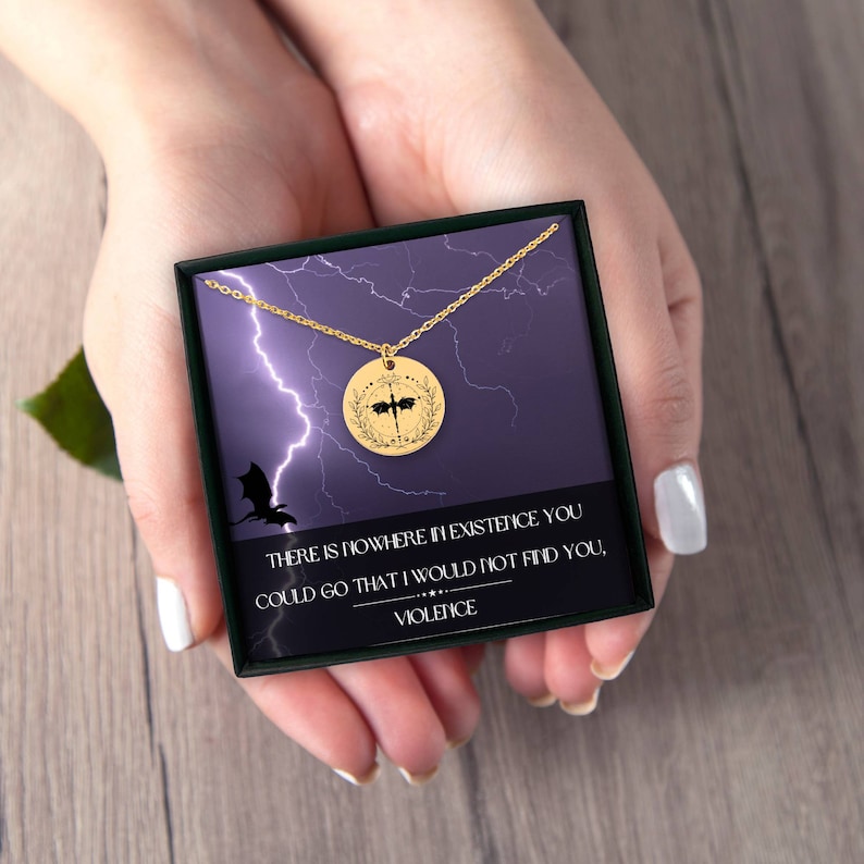 Peut inclure: Un collier en or avec un pendentif rond repr&eacute;sentant une silhouette noire de dragon aux ailes d&eacute;ploy&eacute;es. Le pendentif est entour&eacute; d'une couronne de feuilles. Le collier est dans une bo&icirc;te noire avec un fond violet et des &eacute;clairs. La bo&icirc;te porte le texte "THERE IS NOWHERE IN EXISTENCE YOU COULD GO THAT I WOULD NOT FIND YOU, VIOLENCE".