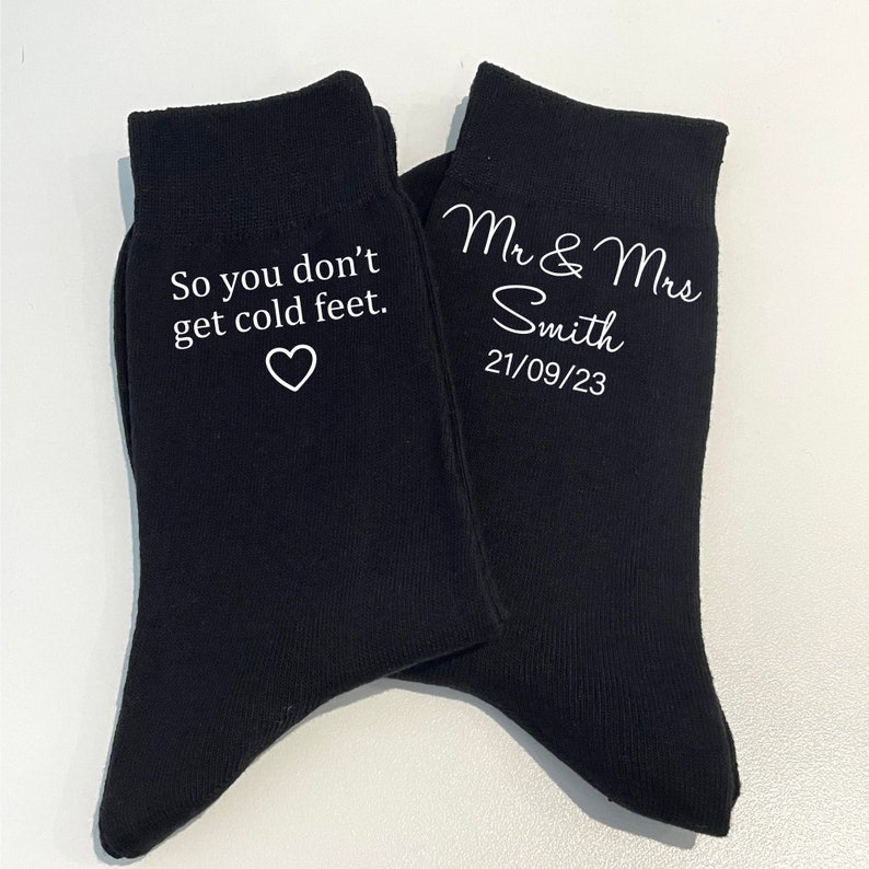 May include: A pair of black socks with white text. One sock says "So you don't get cold feet." with a heart symbol. The other sock says "Mr & Mrs Smith 21/09/23".