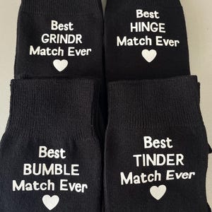 May include: Four pairs of black socks with white text. Each sock reads "Best [dating app name] Match Ever" with a heart symbol. The dating apps featured are Grindr, Hinge, Bumble, and Tinder.