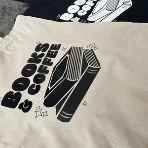 以下が含まれることがあります： 「BOOKS & COFFEE」の文字と、黒と白の積み重ねられた本のイラストが描かれたトートバッグ2つ。1つは白地に黒の文字とイラスト、もう1つは黒地に白の文字とイラストです。