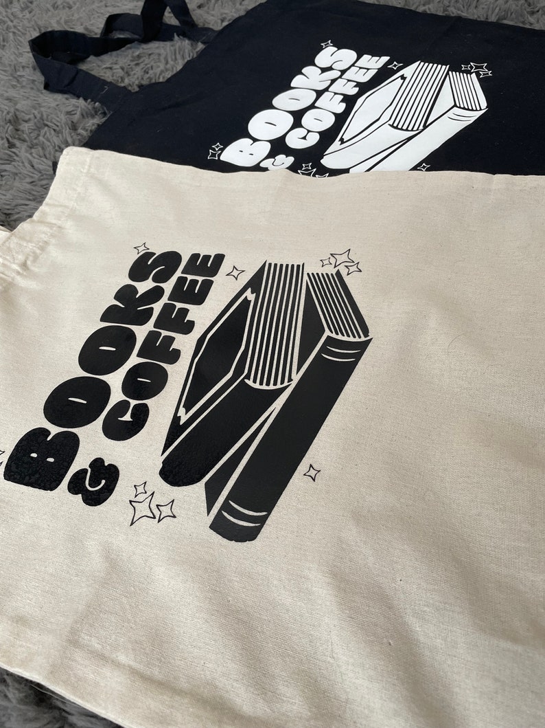 以下が含まれることがあります： 「BOOKS & COFFEE」の文字と、黒と白の積み重ねられた本のイラストが描かれたトートバッグ2つ。1つは白地に黒の文字とイラスト、もう1つは黒地に白の文字とイラストです。