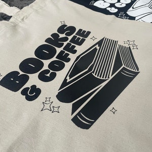 以下が含まれることがあります： 白いキャンバス地のトートバッグに、黒い文字で「BOOKS & COFFEE」と書いてあり、黒い本のシルエットが描かれています。本は背表紙に複数の線があり、テキストと本の周りに小さな星がいくつか散らばっています。