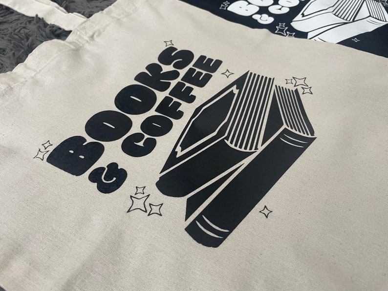 以下が含まれることがあります： 白いキャンバス地のトートバッグに、黒い文字で「BOOKS & COFFEE」と書いてあり、黒い本のシルエットが描かれています。本は背表紙に複数の線があり、テキストと本の周りに小さな星がいくつか散らばっています。