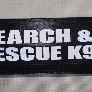 May include: A black rectangular patch with the words "SEARCH & RESCUE K9" in bold white capital letters. The patch is likely made of a durable material and is designed to be attached to a uniform or gear.
