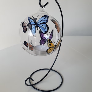 May include: A glass globe candle holder with a black metal stand. The globe is decorated with painted butterflies in shades of blue, purple, yellow, and orange.