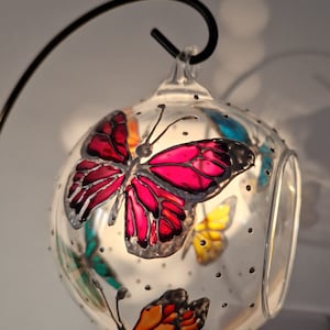 May include: A clear glass globe with a hanging loop, decorated with painted butterflies in shades of pink, orange, yellow, and teal. The globe is filled with small black dots.