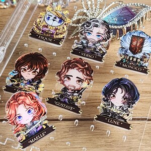 May include: A selection of acrylic charms with anime-style character illustrations. Each charm has a unique design with vibrant colours and is labelled with a name. The charms are on a clear surface.