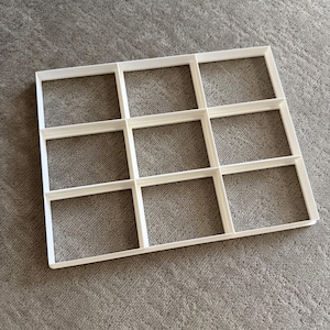 May include: A white, rectangular cookie cutter with nine square openings. The cutter is made of plastic and is designed to cut multiple biscuits or brownies at once. The design is simple and clean, perfect for baking.