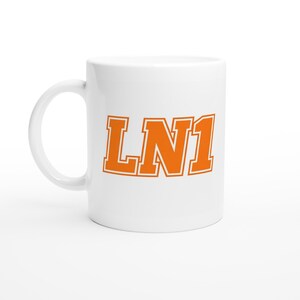 May include: White ceramic mug featuring the orange text "LN1". The mug has a curved handle and a cylindrical shape. The text is in a bold, blocky font with an orange outline.