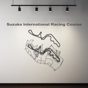 Puede incluir: Arte mural de metal negro que representa el trazado del circuito de Suzuka International Racing Course. El diseño de la pista está montado en una pared blanca, con el texto "Suzuka International Racing Course" encima. La imagen está iluminada por focos.