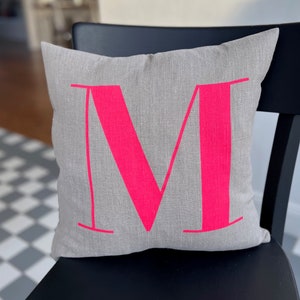 May include: A grey linen cushion with a large, bright pink letter "M" printed on it. The cushion is sitting on a black chair.