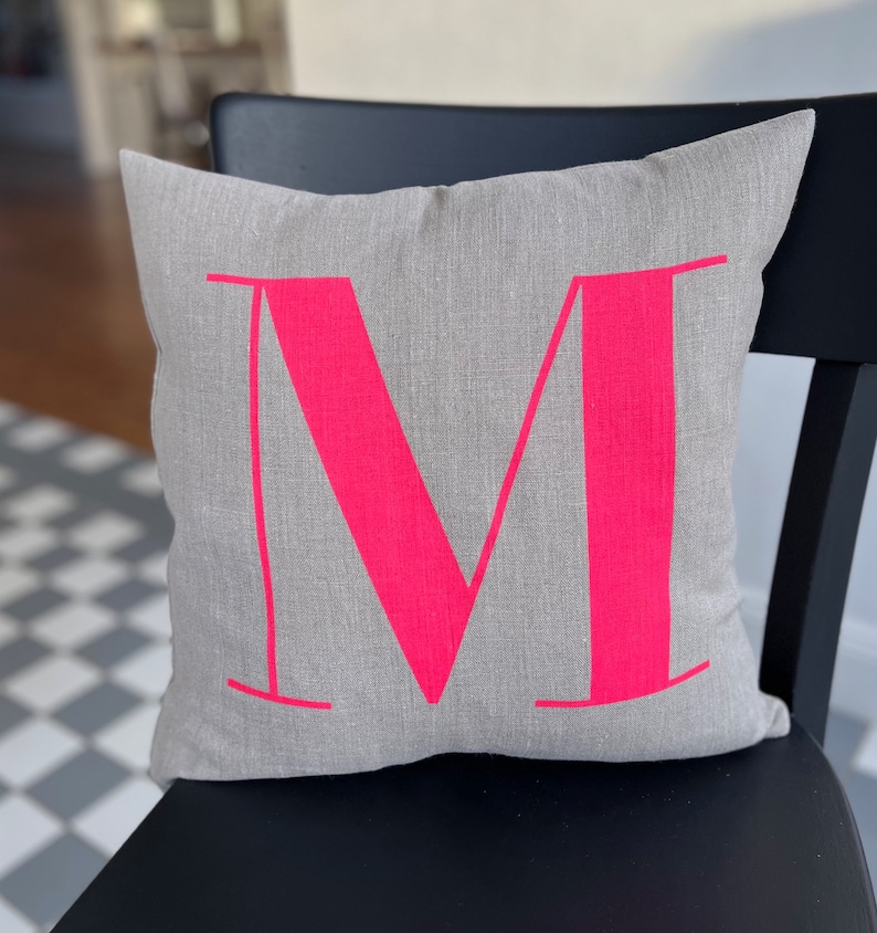 May include: A grey linen cushion with a large, bright pink letter "M" printed on it. The cushion is sitting on a black chair.