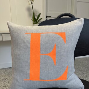 May include: A grey linen cushion with a large orange letter "E" printed on the front.