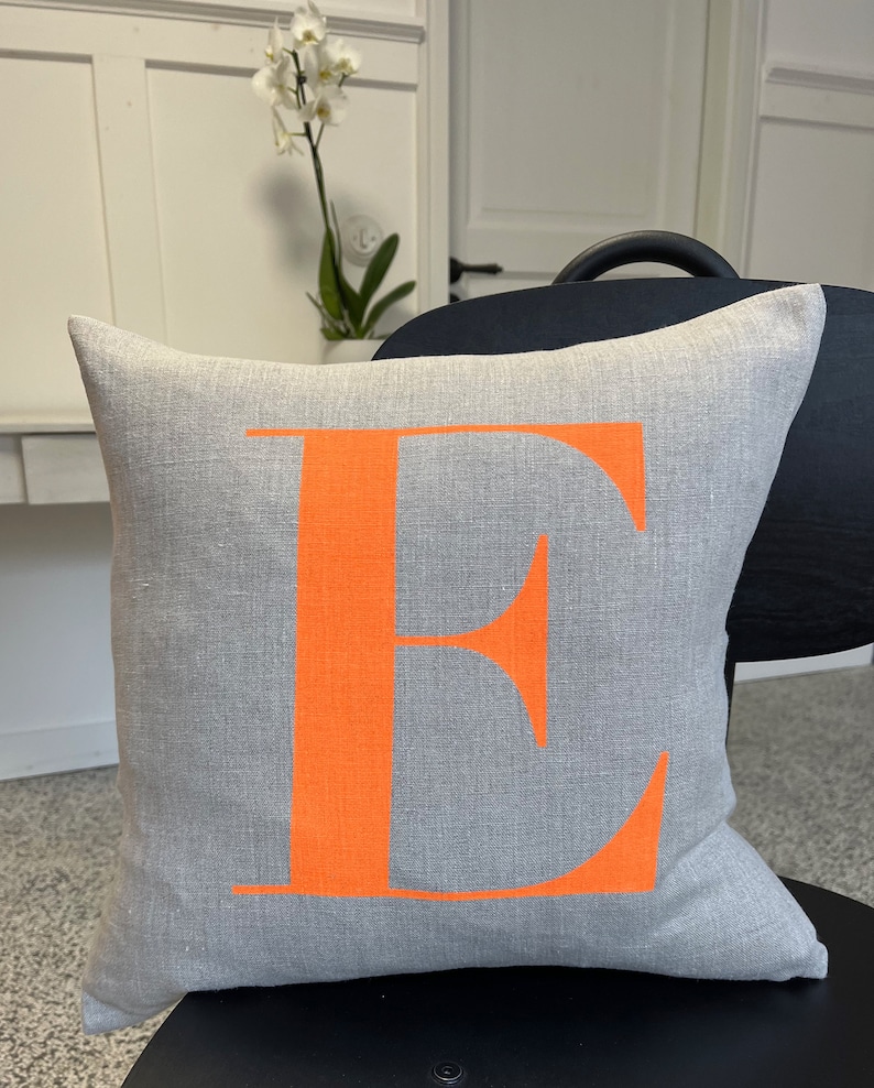 May include: A grey linen cushion with a large orange letter "E" printed on the front.