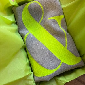 May include: A rectangular grey linen cushion featuring a large neon green ampersand symbol. The cushion is presented in bright green tissue paper, creating a striking contrast. A decorative home accent.