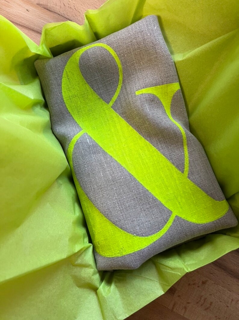 May include: A rectangular grey linen cushion featuring a large neon green ampersand symbol. The cushion is presented in bright green tissue paper, creating a striking contrast. A decorative home accent.