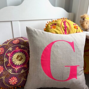 May include: A light grey linen cushion with a large, bright pink letter G printed on the front. The cushion is sitting on a white bench with two other cushions behind it.