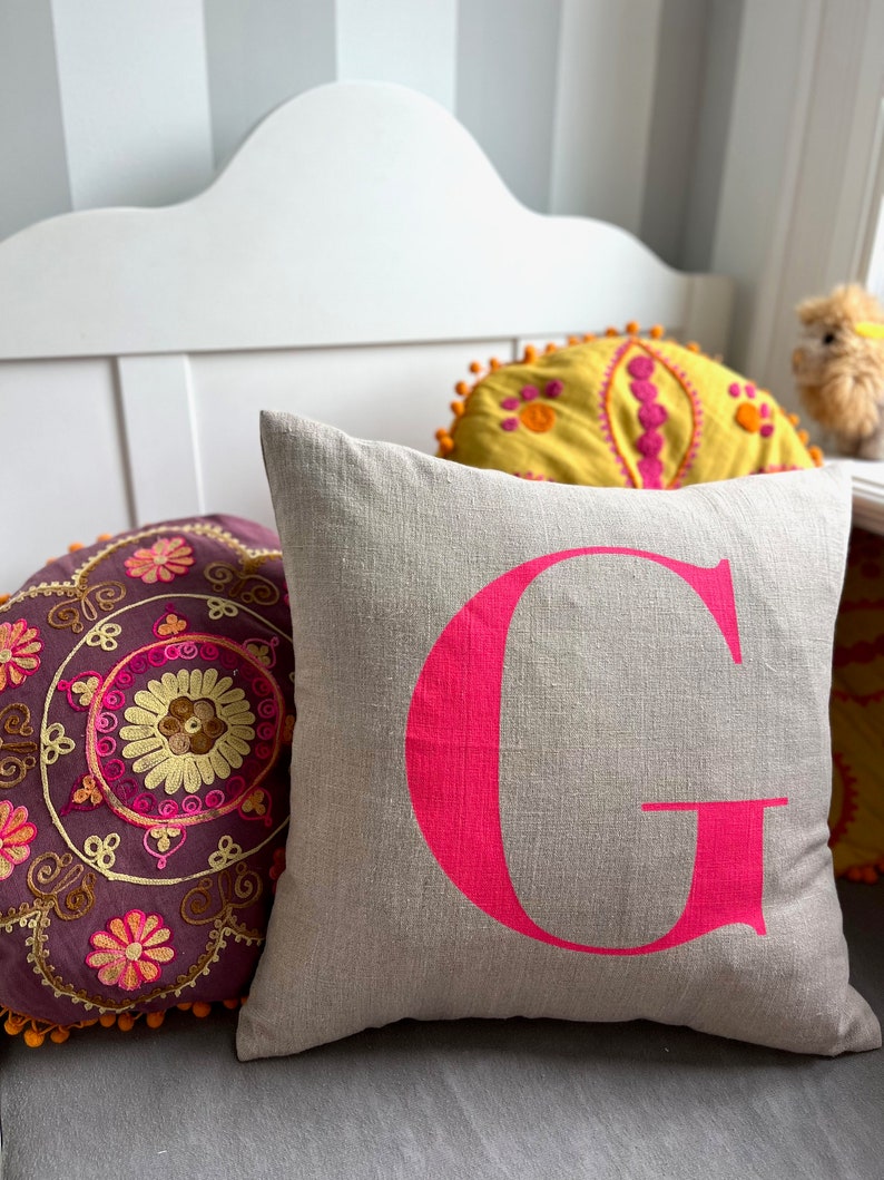 May include: A light grey linen cushion with a large, bright pink letter G printed on the front. The cushion is sitting on a white bench with two other cushions behind it.