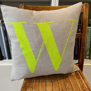 May include: A grey linen cushion with a large, bright green letter "W" printed on the front. The cushion is sitting on a wooden chair.