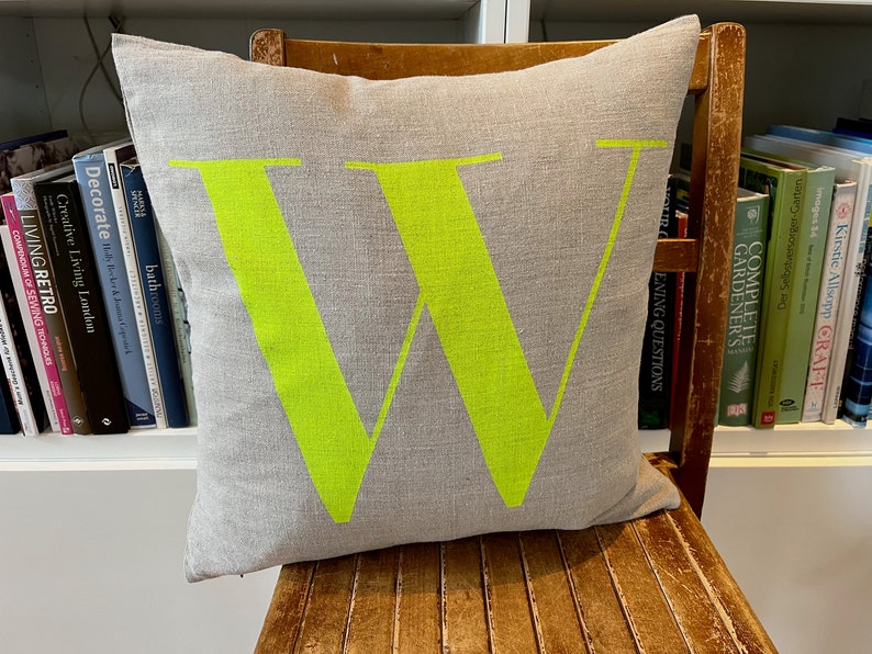 May include: A grey linen cushion with a large, bright green letter "W" printed on the front. The cushion is sitting on a wooden chair.