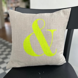 Peut inclure: Un coussin carré décoratif en tissu de type lin neutre. Un grand symbole esperluette vert néon est imprimé sur le devant. Le coussin est posé sur une chaise noire, pour la décoration intérieure.