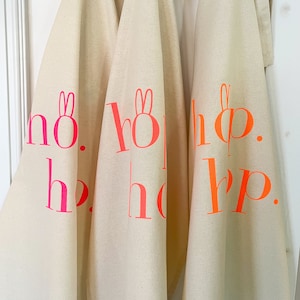 May include: Three white kitchen towels with the words "hop. hop. hop." printed in pink and orange on each towel.