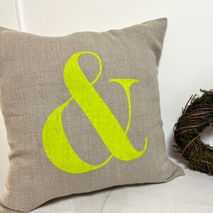 May include: A square, neutral-toned linen cushion featuring a large, bright neon green ampersand symbol. The cushion is on a white surface, with a small wreath in the background. The cushion is made of linen.