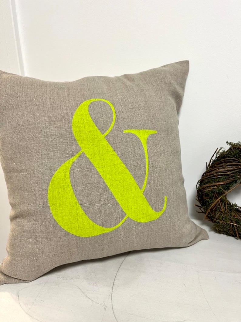 May include: A square, neutral-toned linen cushion featuring a large, bright neon green ampersand symbol. The cushion is on a white surface, with a small wreath in the background. The cushion is made of linen.
