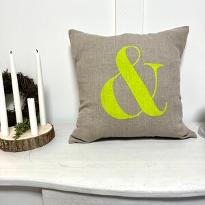 May include: A square, beige cushion featuring a large, vibrant lime green ampersand symbol. The cushion sits on a white table, beside a small wreath and white candles.
