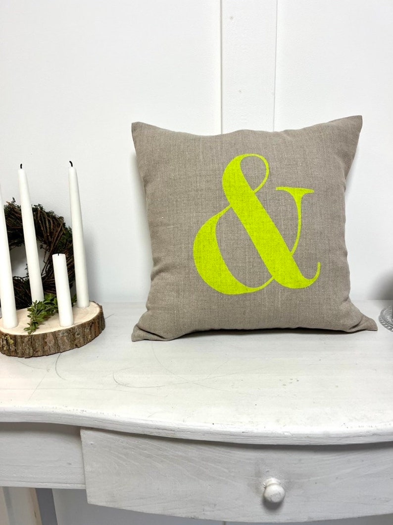 May include: A square, beige cushion featuring a large, vibrant lime green ampersand symbol. The cushion sits on a white table, beside a small wreath and white candles.