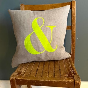May include: A grey cushion featuring a large neon green ampersand symbol. The square cushion sits on a distressed wooden chair. The background is a solid dark blue-grey colour.