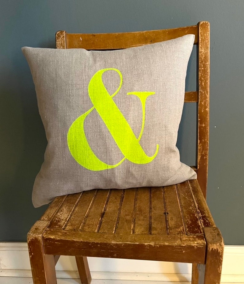 May include: A grey cushion featuring a large neon green ampersand symbol. The square cushion sits on a distressed wooden chair. The background is a solid dark blue-grey colour.