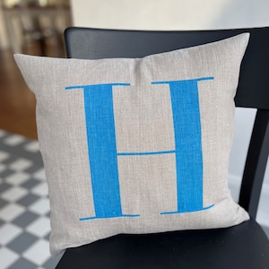 May include: A light grey linen cushion with a large blue letter "H" printed on the front.