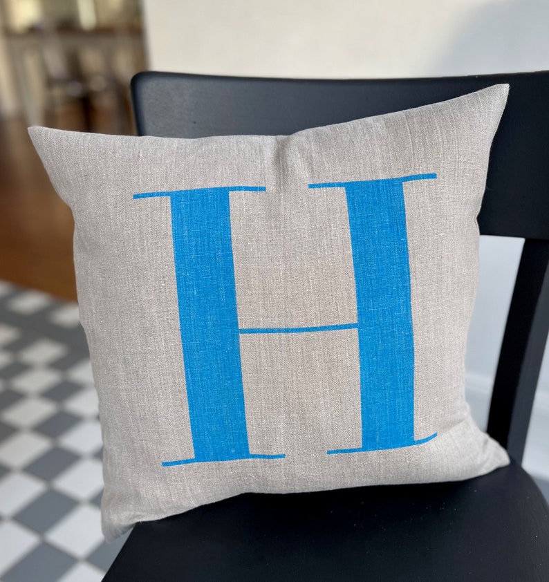 May include: A light grey linen cushion with a large blue letter "H" printed on the front.