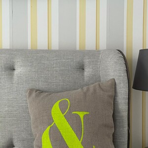 May include: A brown cushion with a neon green ampersand symbol, placed on a grey upholstered headboard. The background features striped wallpaper in white, yellow, and grey. The image also shows framed artwork.